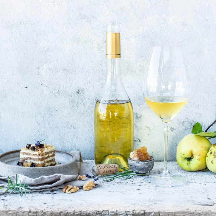 Eine Flasche süßer Wein mit goldenem Korken, ein großes Weinglas mit gelbem Wein, ein Stück Schichtkuchen, eine kleine Schale mit kandiertem Obst, Walnüsse und grüne Äpfel.