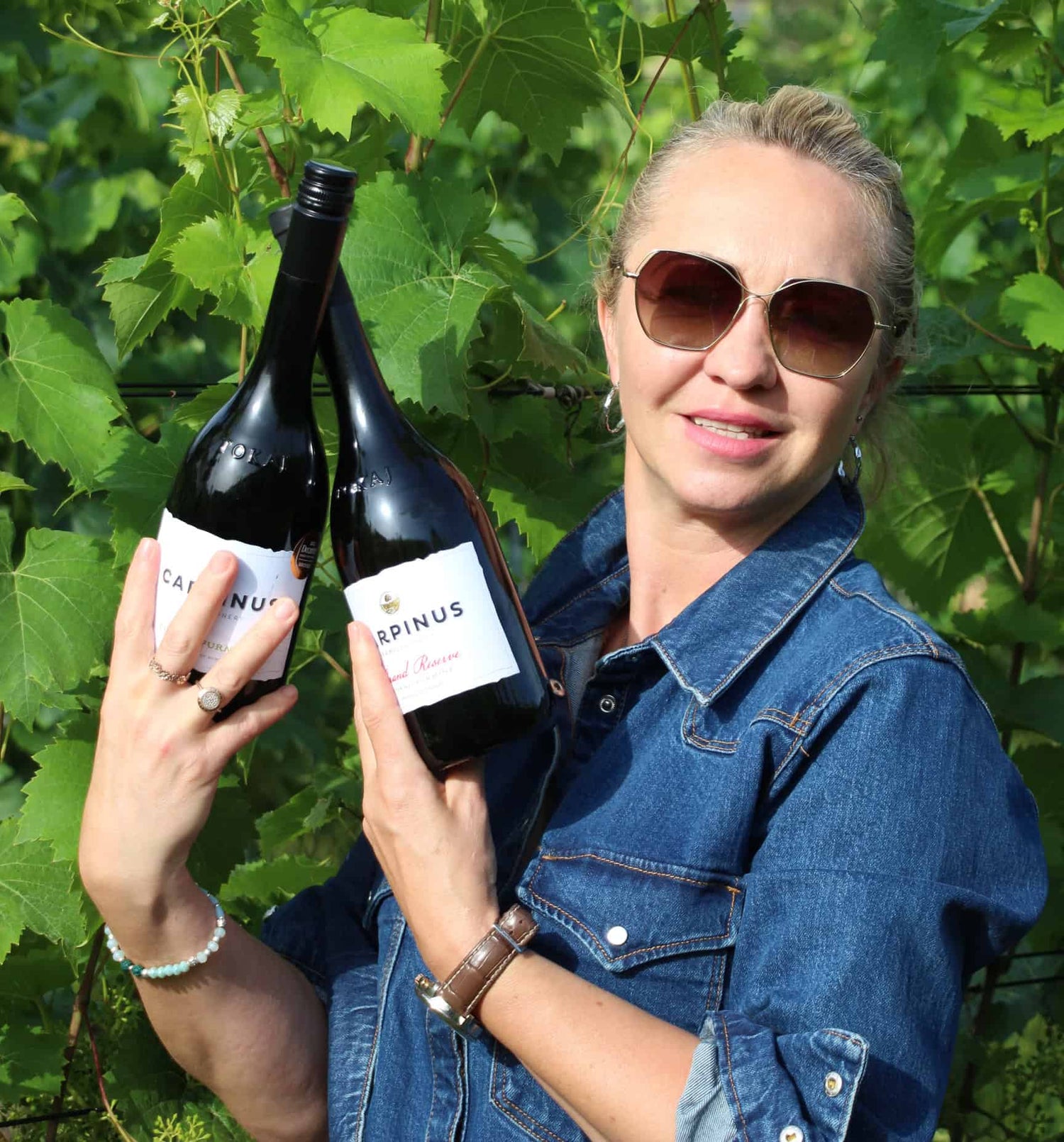 Frau hält zwei Flaschen Wein mit dem Etikett 'CARPINUS' in den Händen. Sie trägt eine Jeansjacke und hat eine Armbanduhr am Handgelenk.