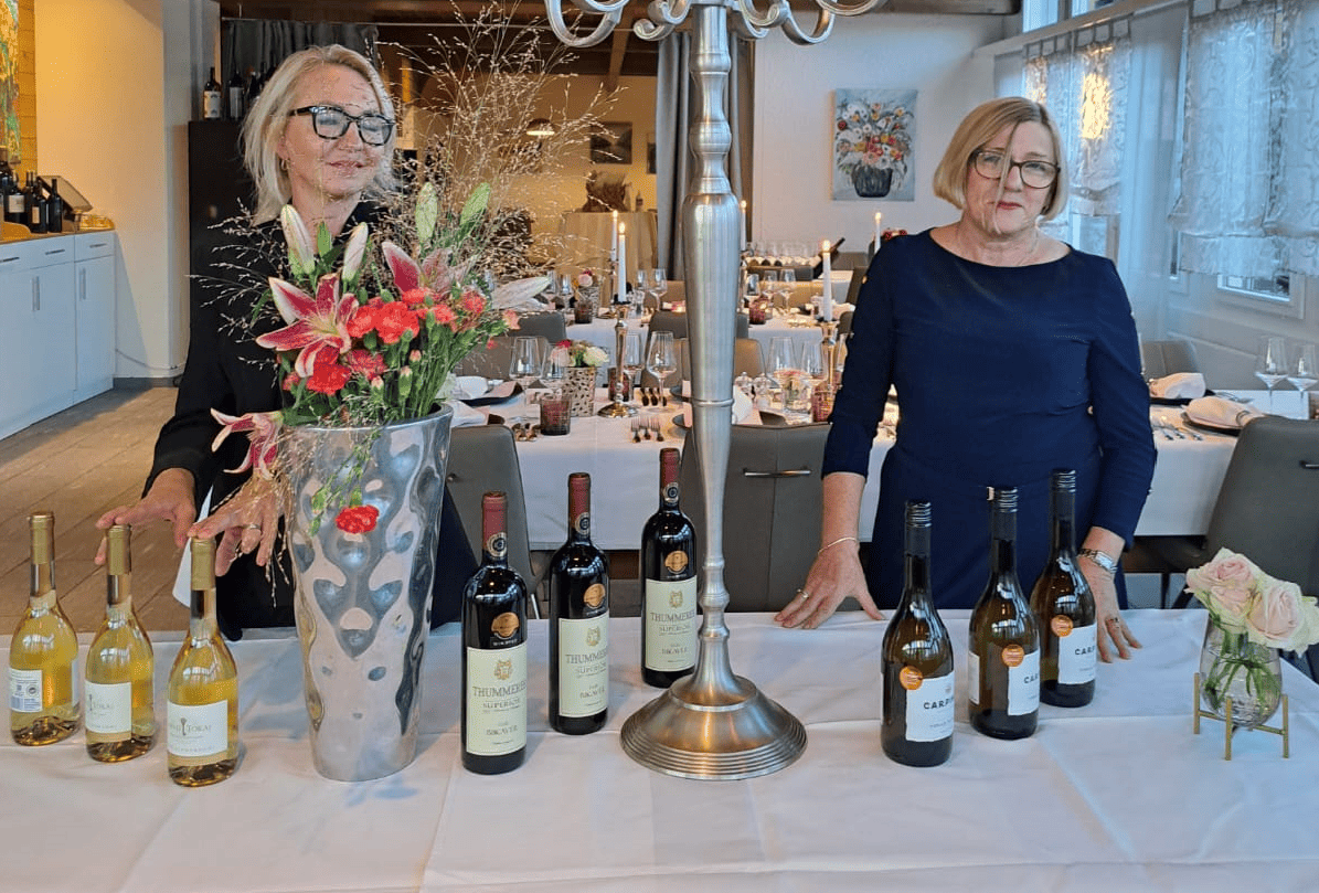 Zwei Frauen stehen an einem Tisch mit einer Vase voller Blumen und mehreren Weinflaschen. Links sind drei Flaschen Weißwein und vier Flaschen Rotwein aufgereiht.