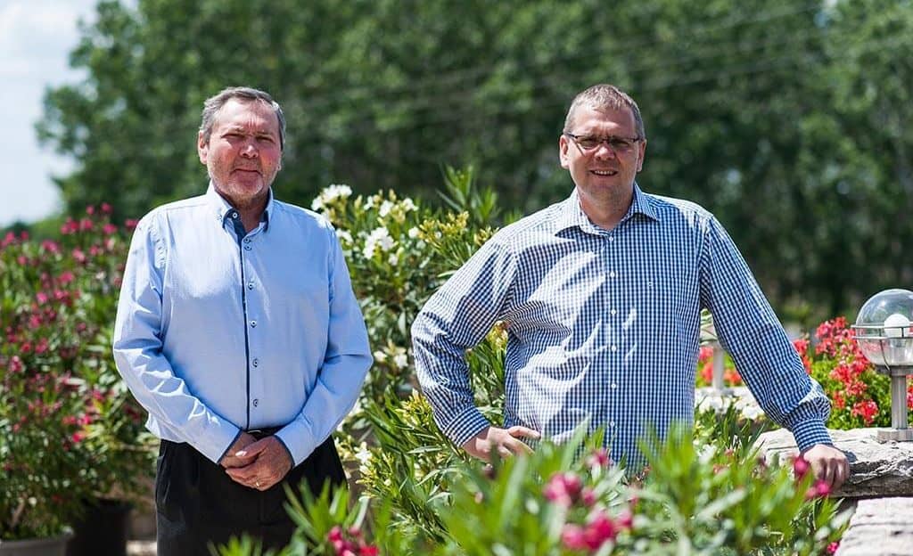 Zwei Männer stehen in einem Garten mit blühenden Pflanzen. Der Mann links trägt ein hellblaues Hemd, der Mann rechts ein kariertes Hemd. Im Hintergrund sind Bäume sichtbar.
