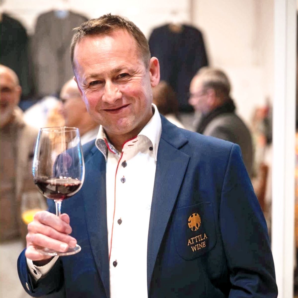 Mann in einem blauen Blazer mit dem Logo 'ATILLA WINE', hält ein Weinglas mit rotem Wein in der rechten Hand. Er trägt ein weißes Hemd.