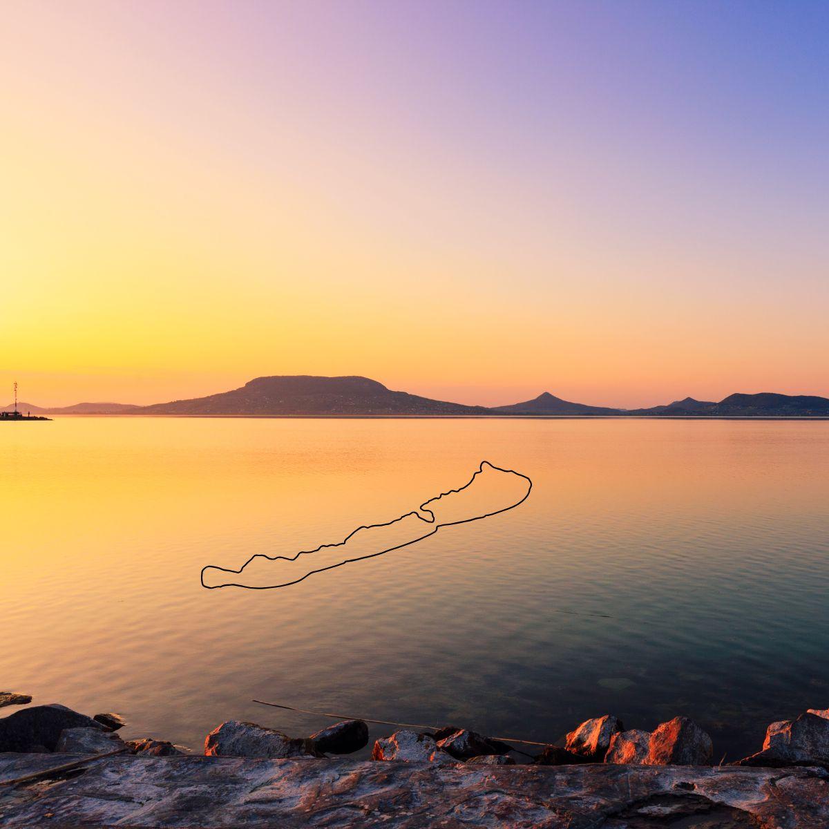 Sonnenuntergang über dem Balaton mit ruhigem Wasser und silhouettierten Bergen im Hintergrund. Steine im Vordergrund.