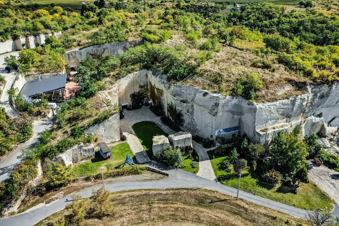 Luftaufnahme eines Steinbruchs mit steilen, weißen Felswänden, umgeben von Bäumen und Sträuchern. Ein gepflasterter Weg führt durch die Landschaft.