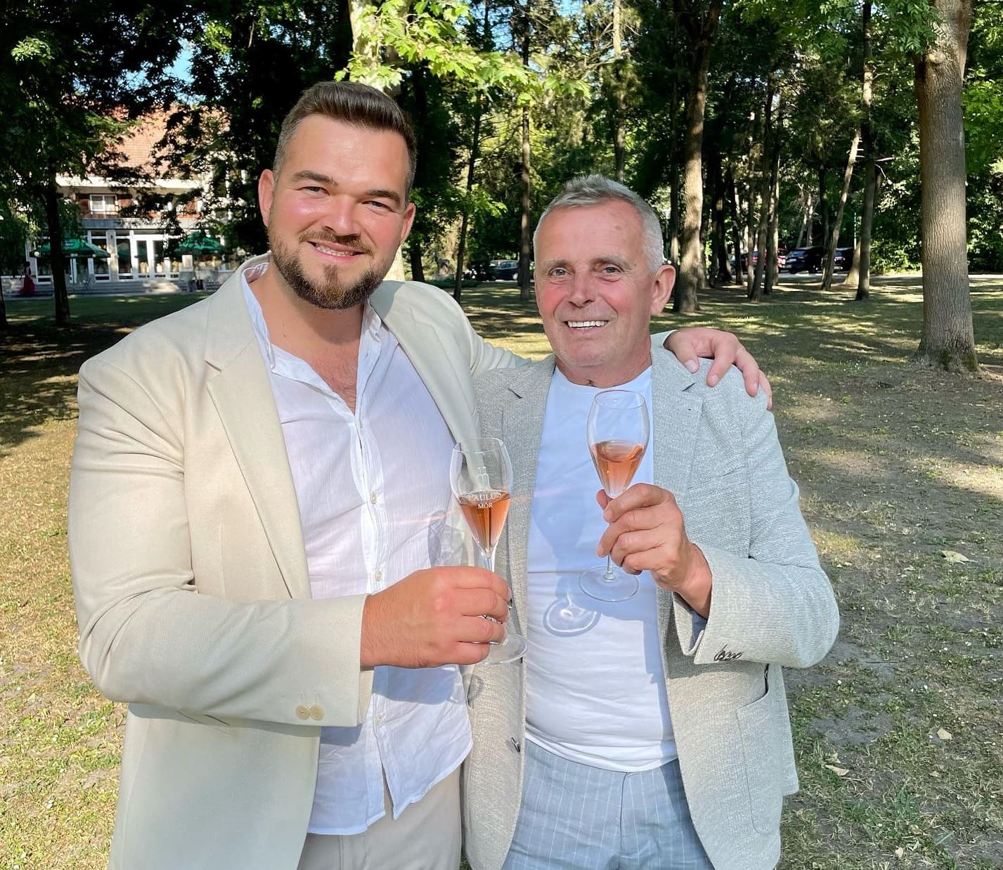 Zwei Männer stehen im Freien und halten jeweils ein Glas mit roséfarbenem Getränk. Der Mann links trägt einen hellen Anzug und ein weißes Hemd, der Mann rechts einen hellen Blazer über einem weißen T-Shirt.