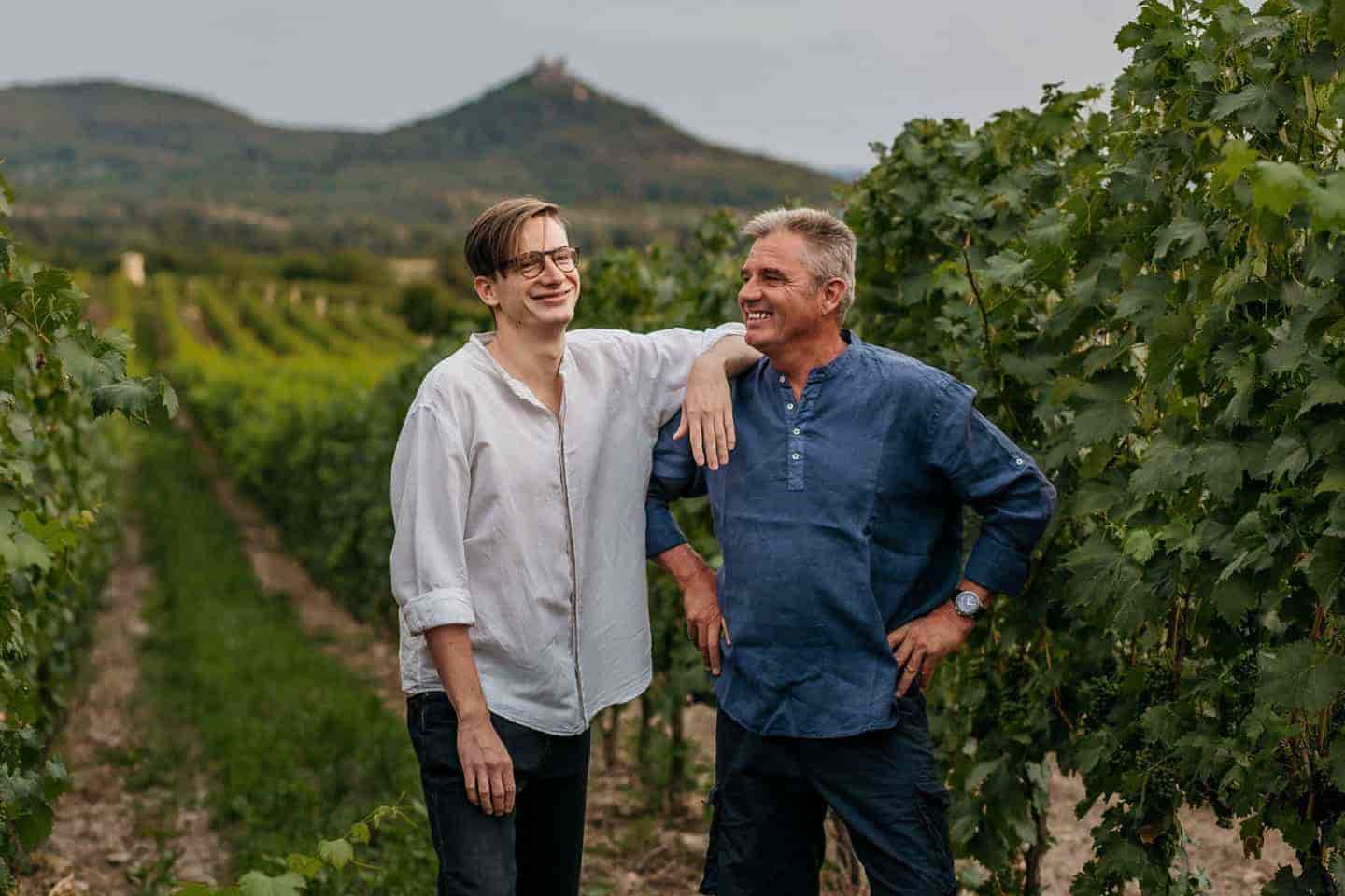 Zwei Personen stehen in einem Weinberg zwischen Reben. Die linke Person trägt ein weißes Hemd, die rechte ein blaues Hemd. Im Hintergrund ist ein Hügel mit einer Burg sichtbar.