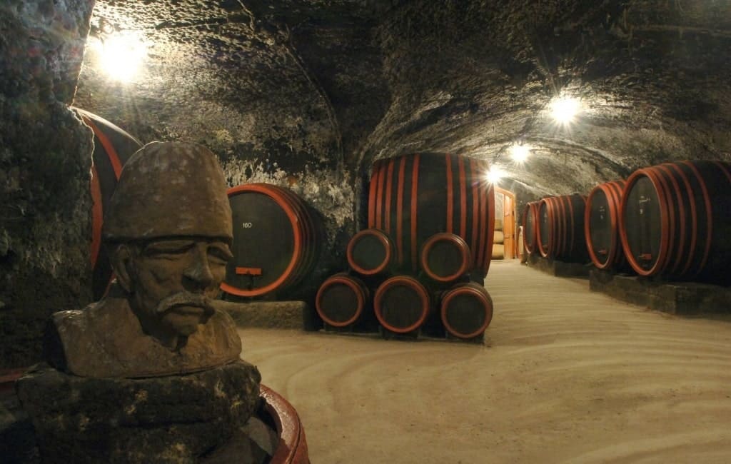 Ein Weinkeller mit runden, großen Fässern in Schwarz und Rot, die an der Wand stehen. Im Vordergrund eine Statue auf einem Sockel. Decke mit Beleuchtung.