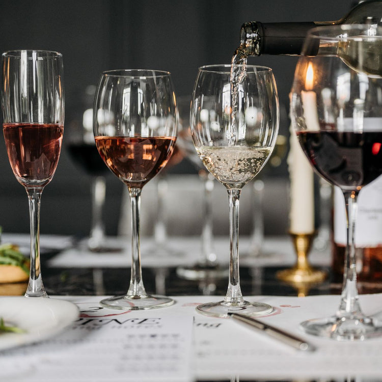 Drei Weingläser mit Rosé, Weiß- und Rotwein auf einem Tisch. Eine Flasche wird in ein Glas mit sprudelndem Weißwein gegossen. Im Hintergrund sind weitere Gläser sichtbar.