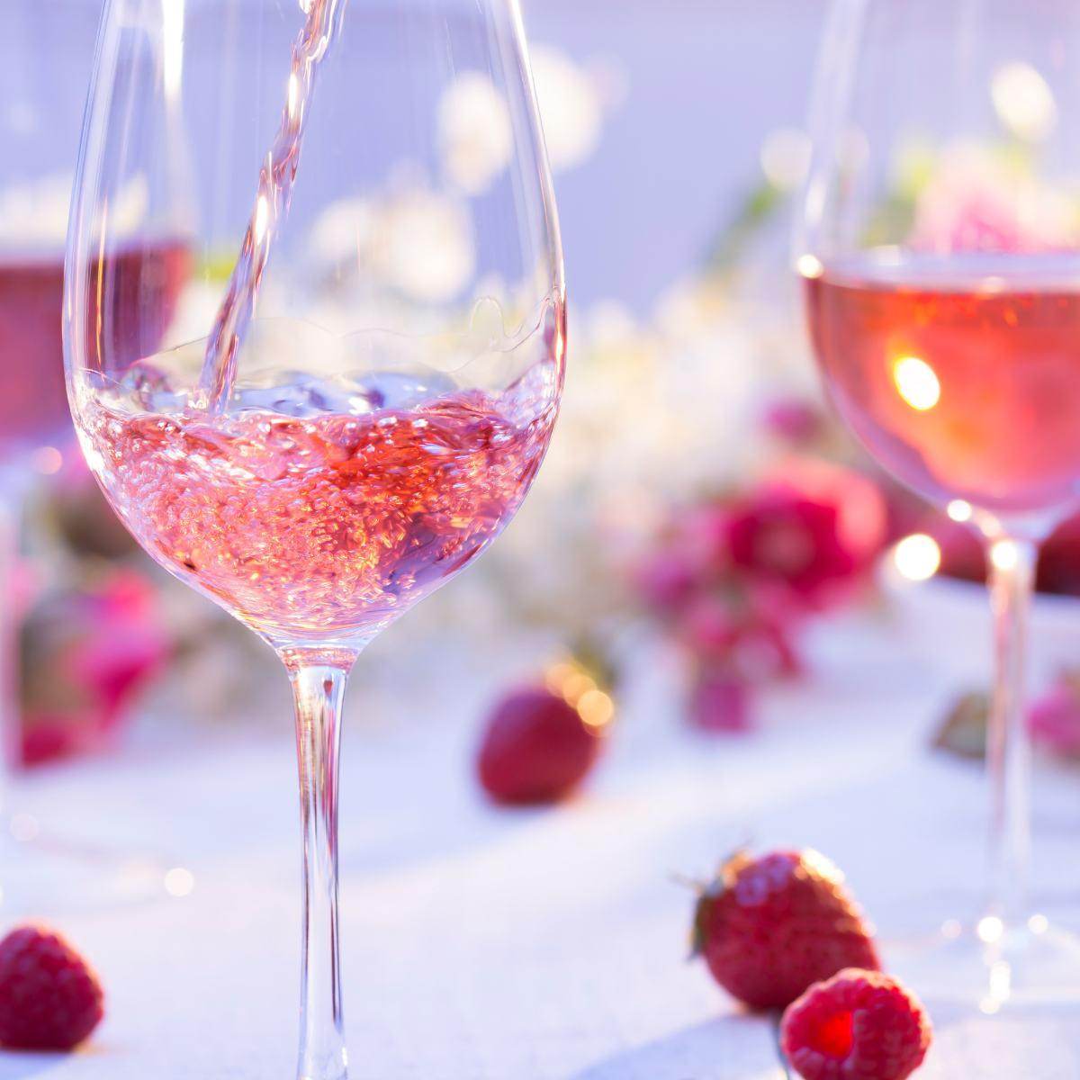 Ein Glas mit roséfarbenem Wein wird gefüllt, während ein weiteres Glas im Hintergrund sichtbar ist. Um das Glas liegen frische Erdbeeren und Himbeeren.