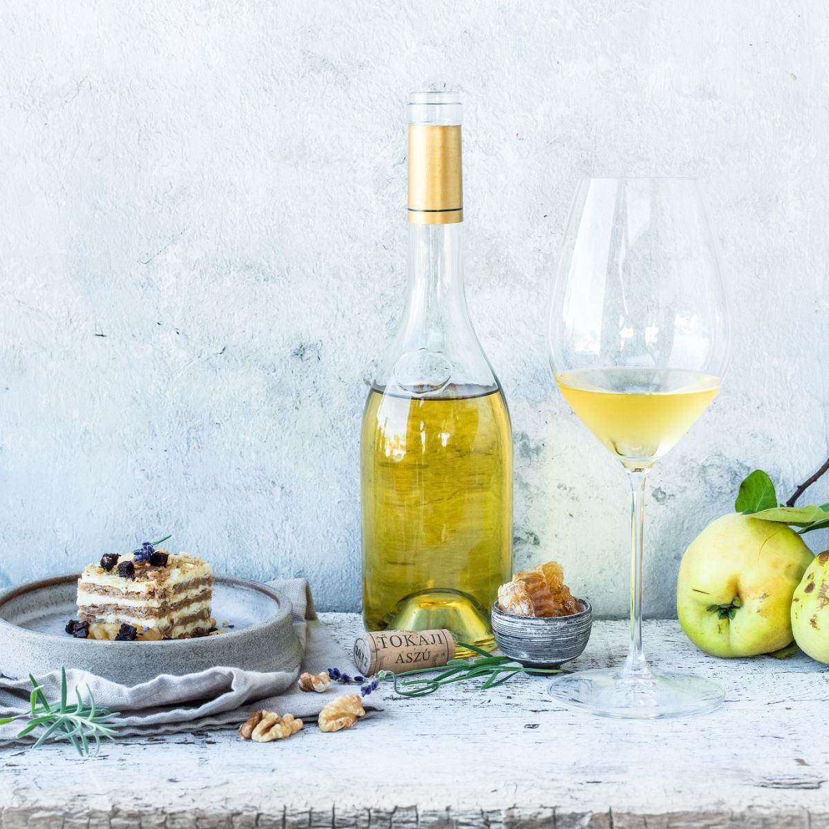 Eine Flasche süßer Wein mit goldenem Korken, ein großes Weinglas mit gelbem Wein, ein Stück Schichtkuchen, eine kleine Schale mit kandiertem Obst, Walnüsse und grüne Äpfel.