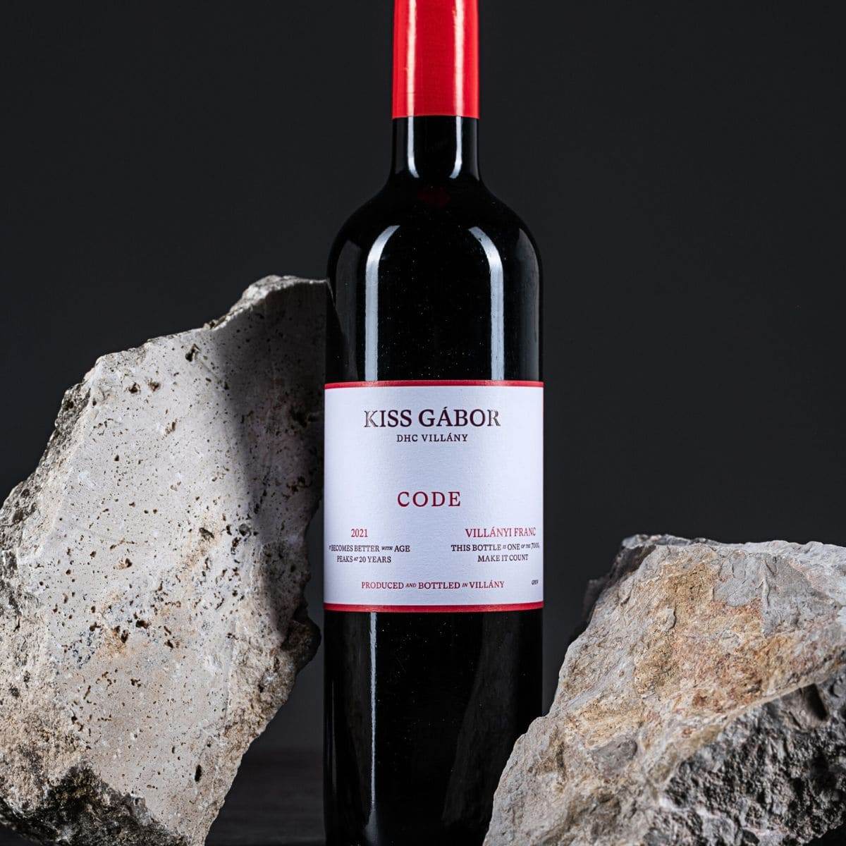 Flasche Kiss Gábor Code Rotwein mit rotem Verschluss zwischen zwei großen grauen Steinen vor dunklem Hintergrund