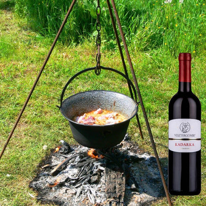Schwarzer Kessel mit kochendem Essen hängt über offenem Feuer auf einer Wiese, daneben Flasche Vesztergombi Kadarka Rotwein