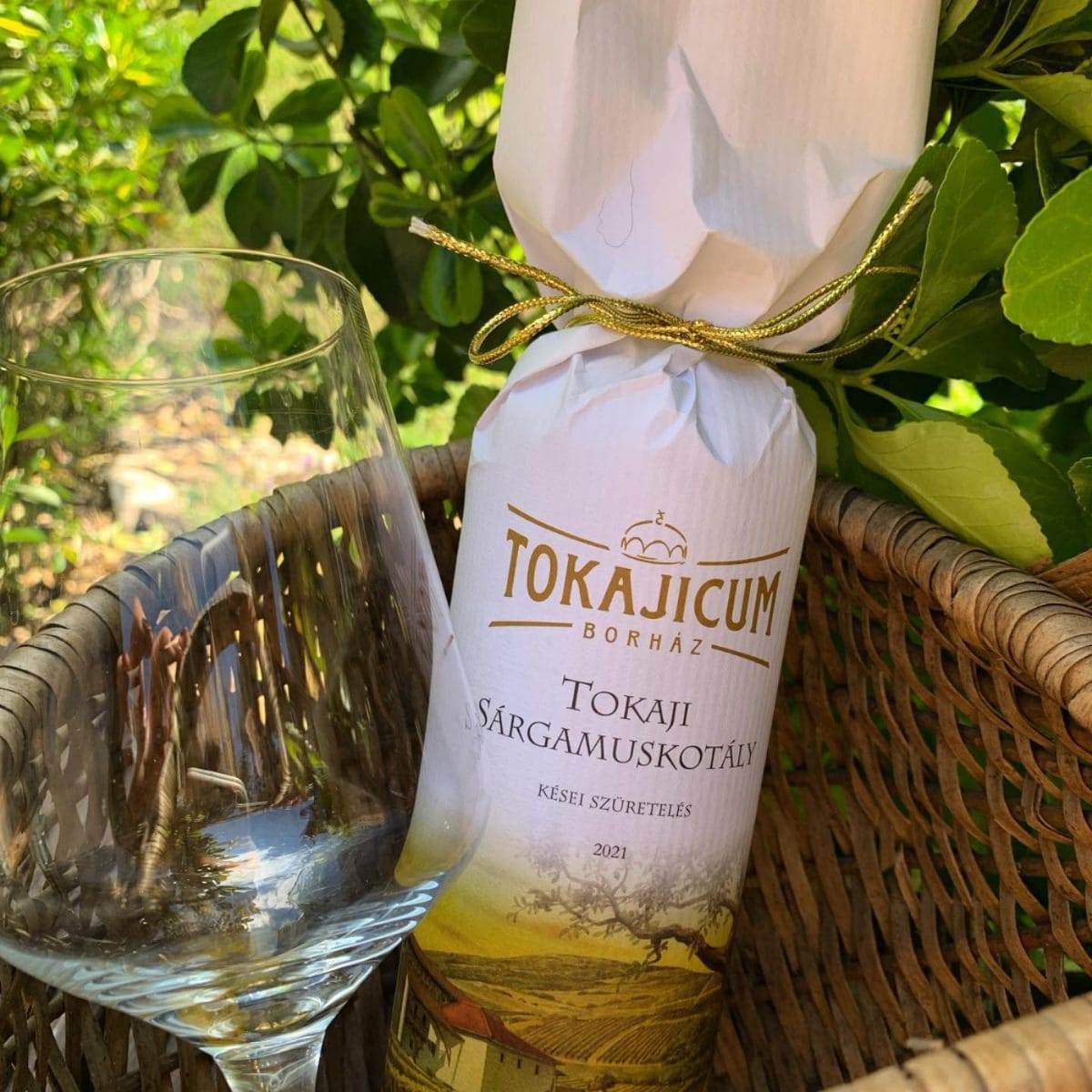 Flasche Tokajicum Tokaji Sárgamuskotály 2021 Wein in weißem Papier mit goldener Schnur neben leerem Weinglas im Korb