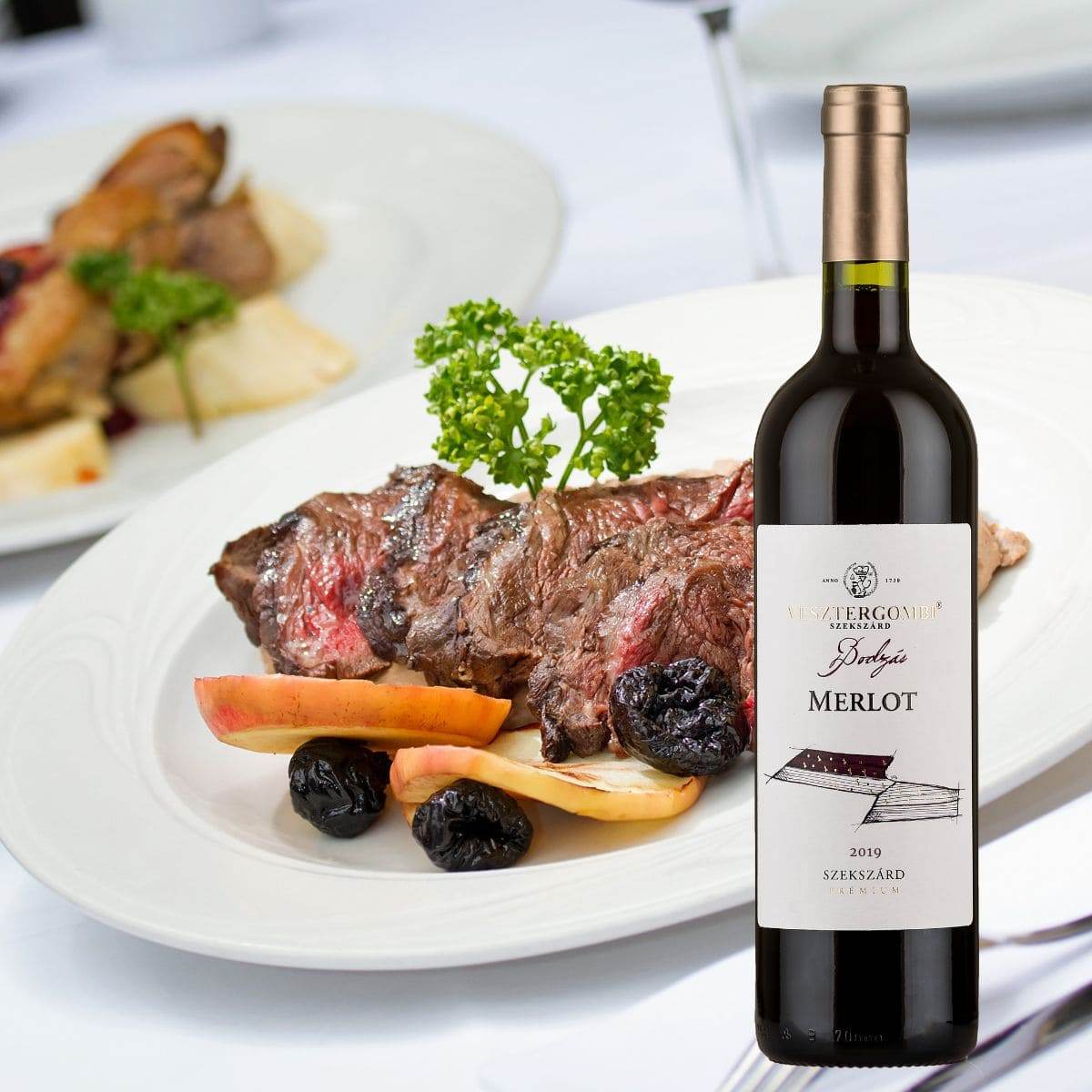 Gegrilltes Steak mit Apfelscheiben und getrockneten Pflaumen auf weißem Teller, daneben Flasche Vesztergombi Bodzás Merlot