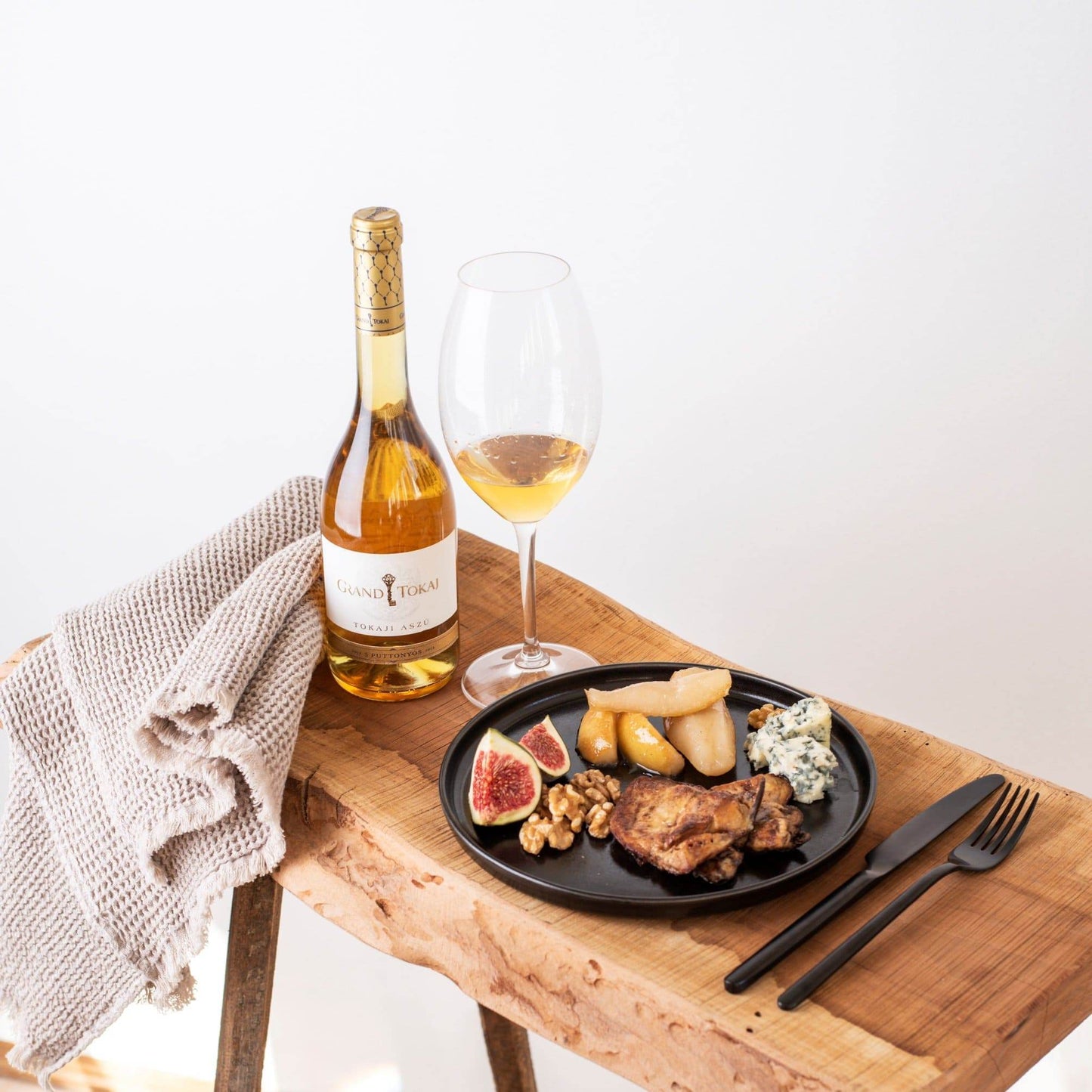 Holztisch mit einer Flasche Grand Tokaj Wein, einem Glas mit weißem Wein, einem schwarzen Teller mit Feigen, Walnüssen, Birne