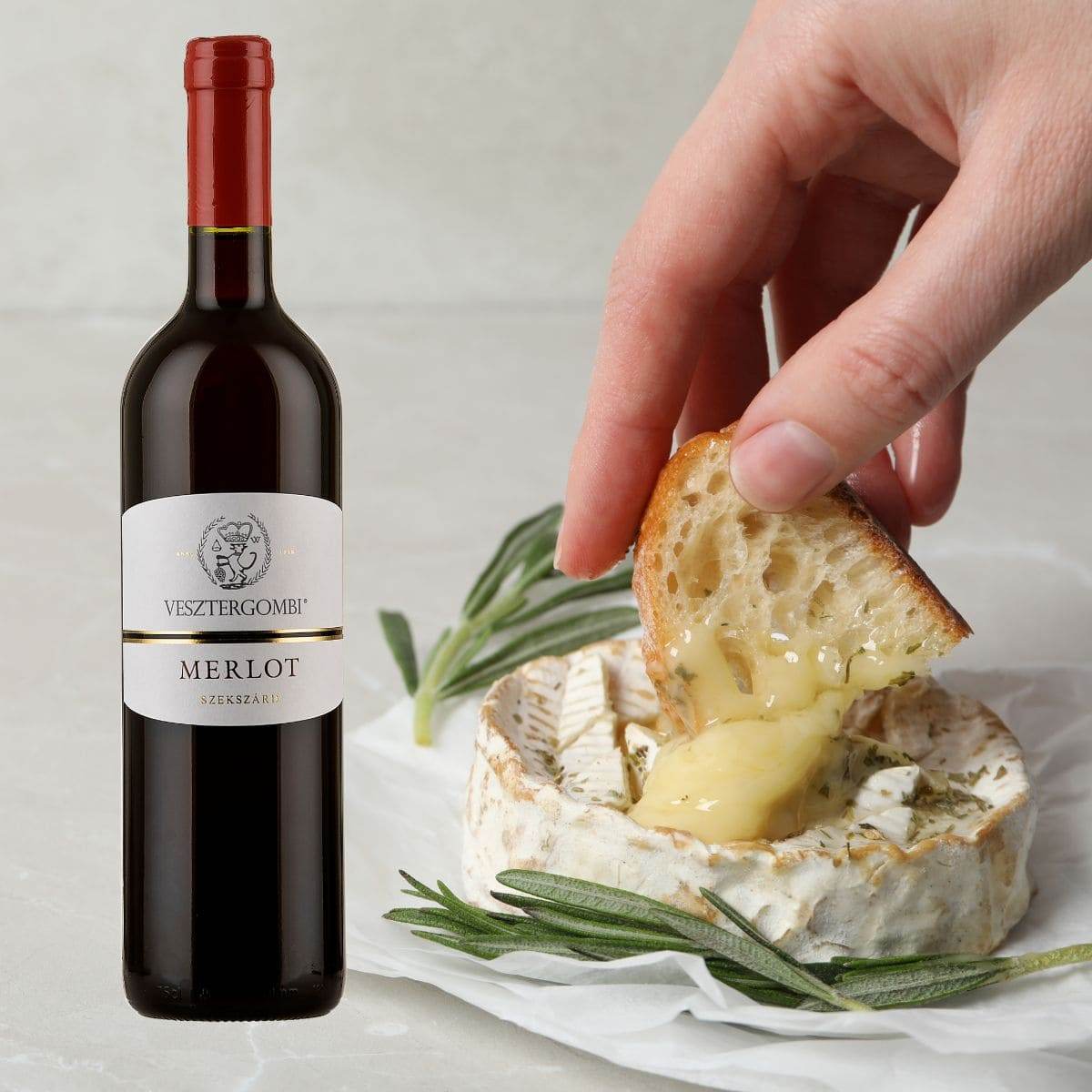 Flasche Vesztergombi Merlot Rotwein neben geschmolzenem Camembert mit Rosmarin und Hand, die ein Stück Brot eintaucht