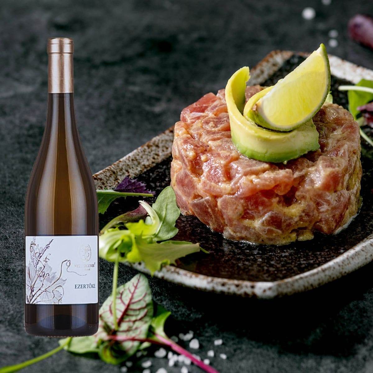 Runde Portion roher Fisch-Tatar mit Avocado & Limettenscheibe auf schwarzem Teller, daneben Flasche Wein mit Etikett Ezertőke