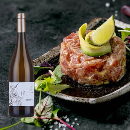 Runde Portion roher Fisch-Tatar mit Avocado & Limettenscheibe auf schwarzem Teller, daneben Flasche Wein mit Etikett Ezertőke