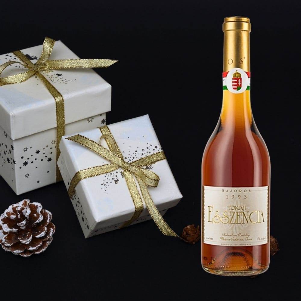 Flasche Majoros Tokaji Esszencia 1993 mit zwei weißen Geschenkboxen mit goldenen Schleifen und zwei Kiefernzapfen auf schwarz