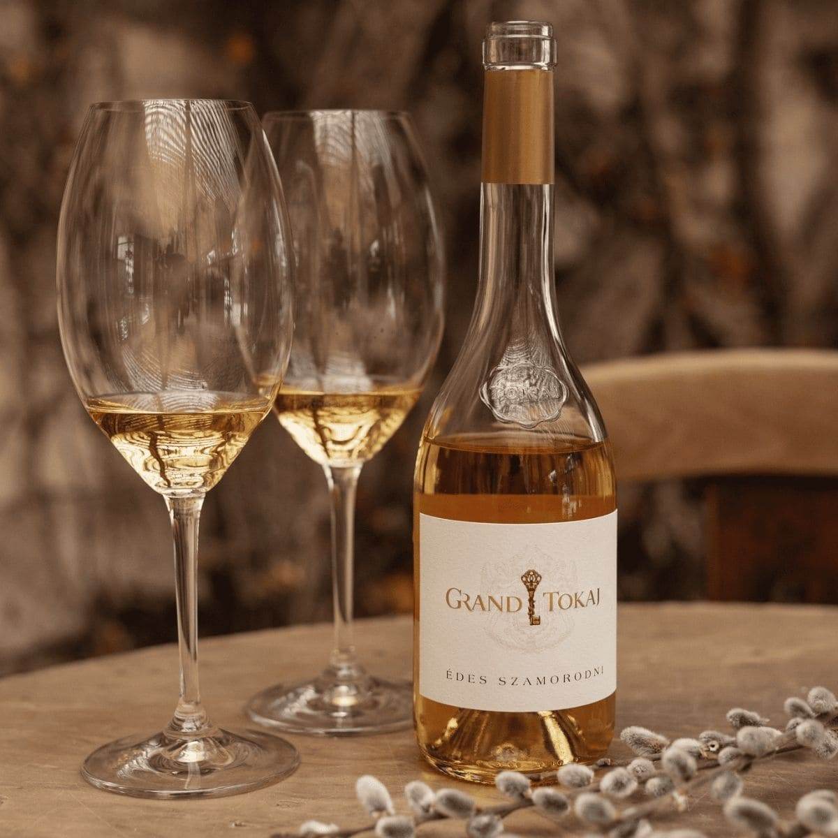 Flasche Grand Tokaj Édes Szamorodni Weisswein und zwei gefüllte Weingläser auf einem Holztisch mit Weidenkätzchen
