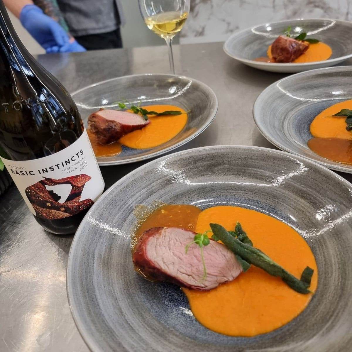 Vier Teller mit rosa gebratenem Fleisch auf orangener Sauce, garniert mit grünen Kräutern, daneben eine Flasche Basic Instincts.