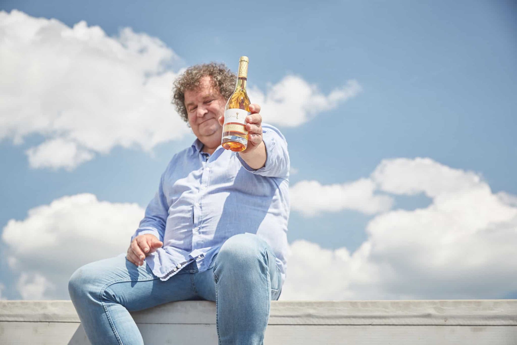 Mann mit lockigem Haar sitzt auf einer Mauer und hält eine Flasche Aszúwein in der Hand. Himmel mit Wolken im Hintergrund.