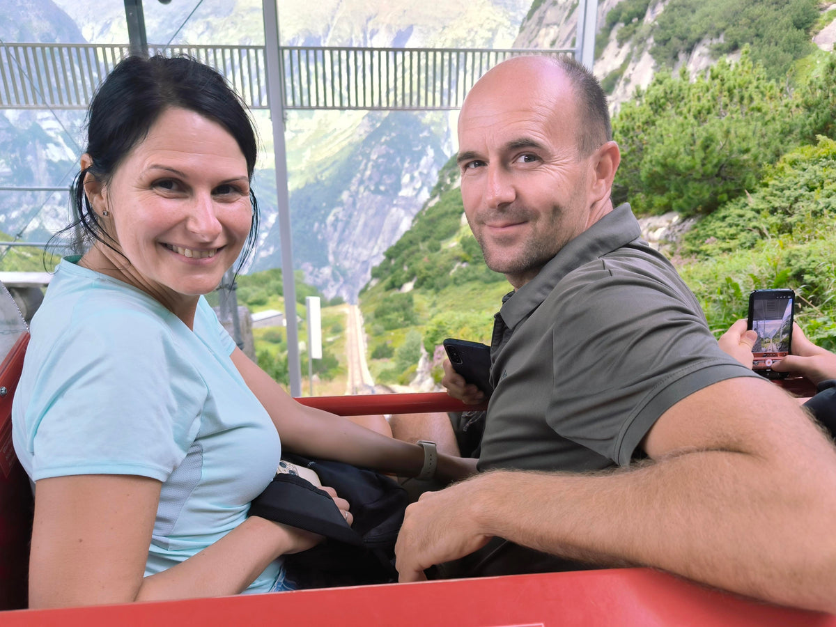 Zwei Menschen sitzen nebeneinander in einer roten Seilbahngondel und lächeln in die Kamera, im Hintergrund sind Berge und grüne Vegetation sichtbar.