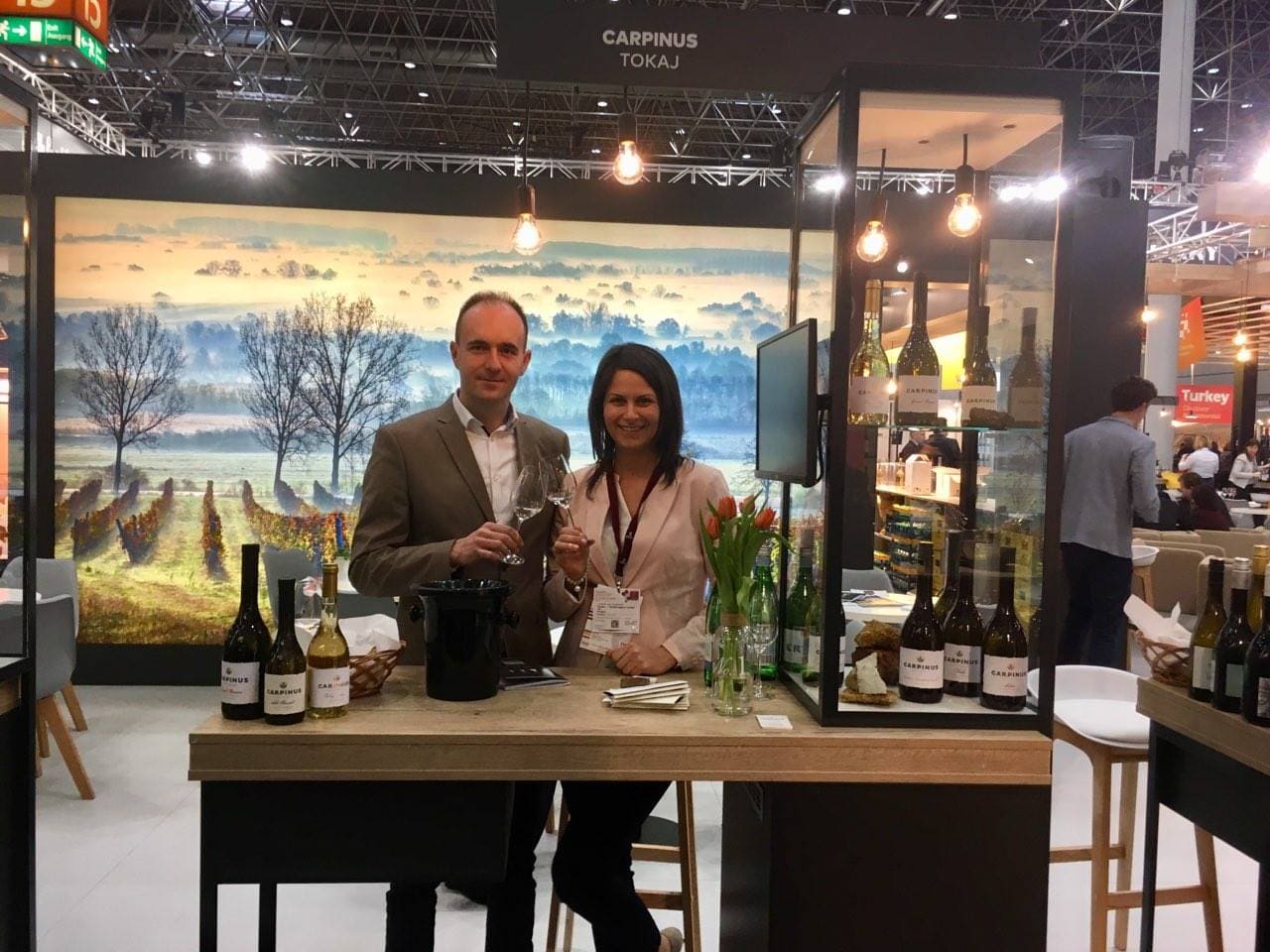Zwei Personen stehen an einem Stehtisch in einem Messestand. Eine Person hält ein Glas, die andere eine Flasche Wein. Im Hintergrund ist eine Landschaft mit Weinreben und Bäumen zu sehen. Auf dem Tisch stehen mehrere Weinflaschen und eine Vase mit Tulpen.