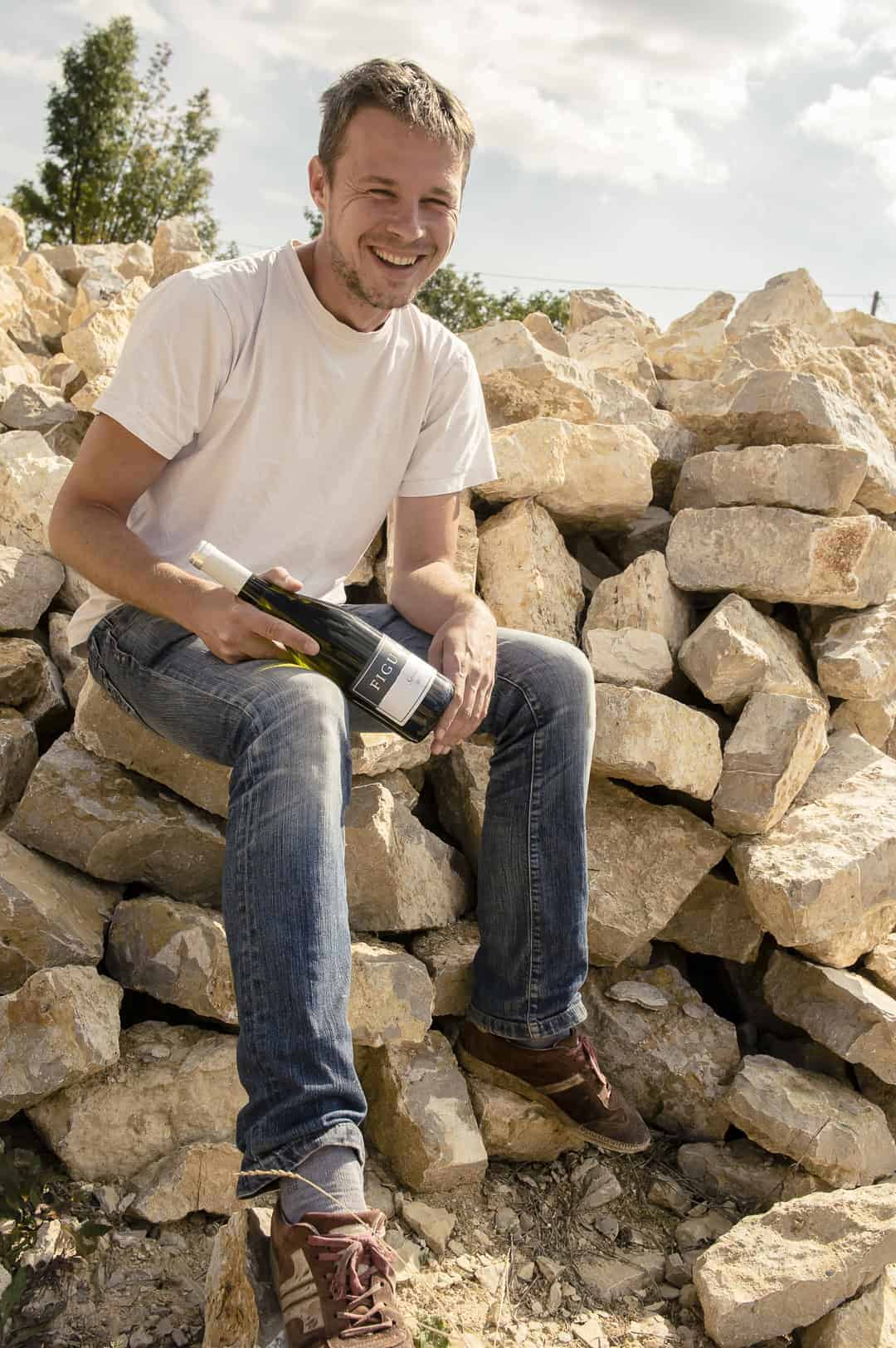 Junger Mann in weißem T-Shirt und blauen Jeans sitzt auf einem Steinhaufen und hält eine Flasche Wein in der Hand.