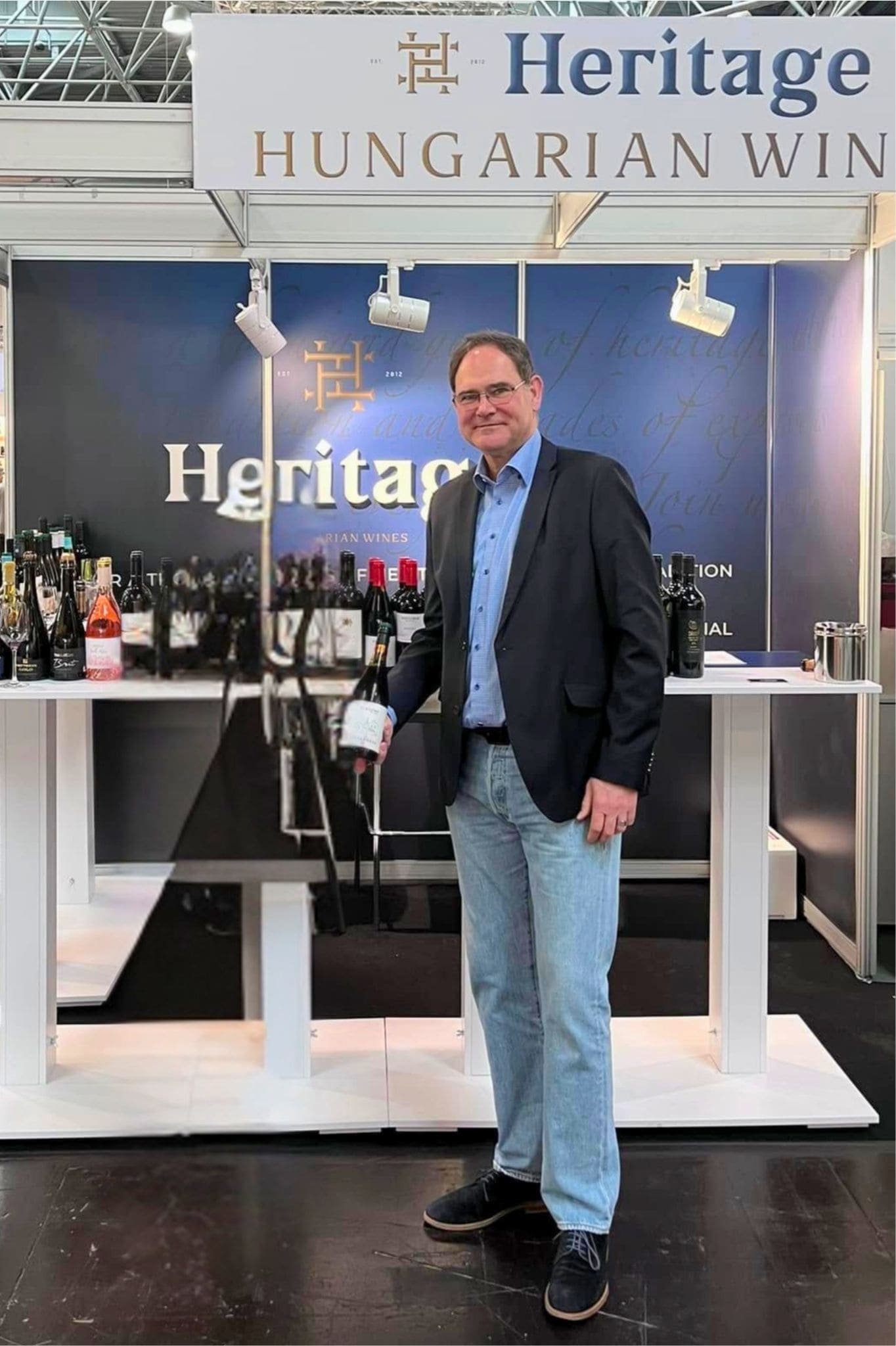 Mann in schwarzem Blazer und blauer Hemd steht vor einem Stand mit Weinen. Auf dem Stand sind Flaschen und Gläser sichtbar. Oben steht 'Heritage HUNGARIAN WINE'.