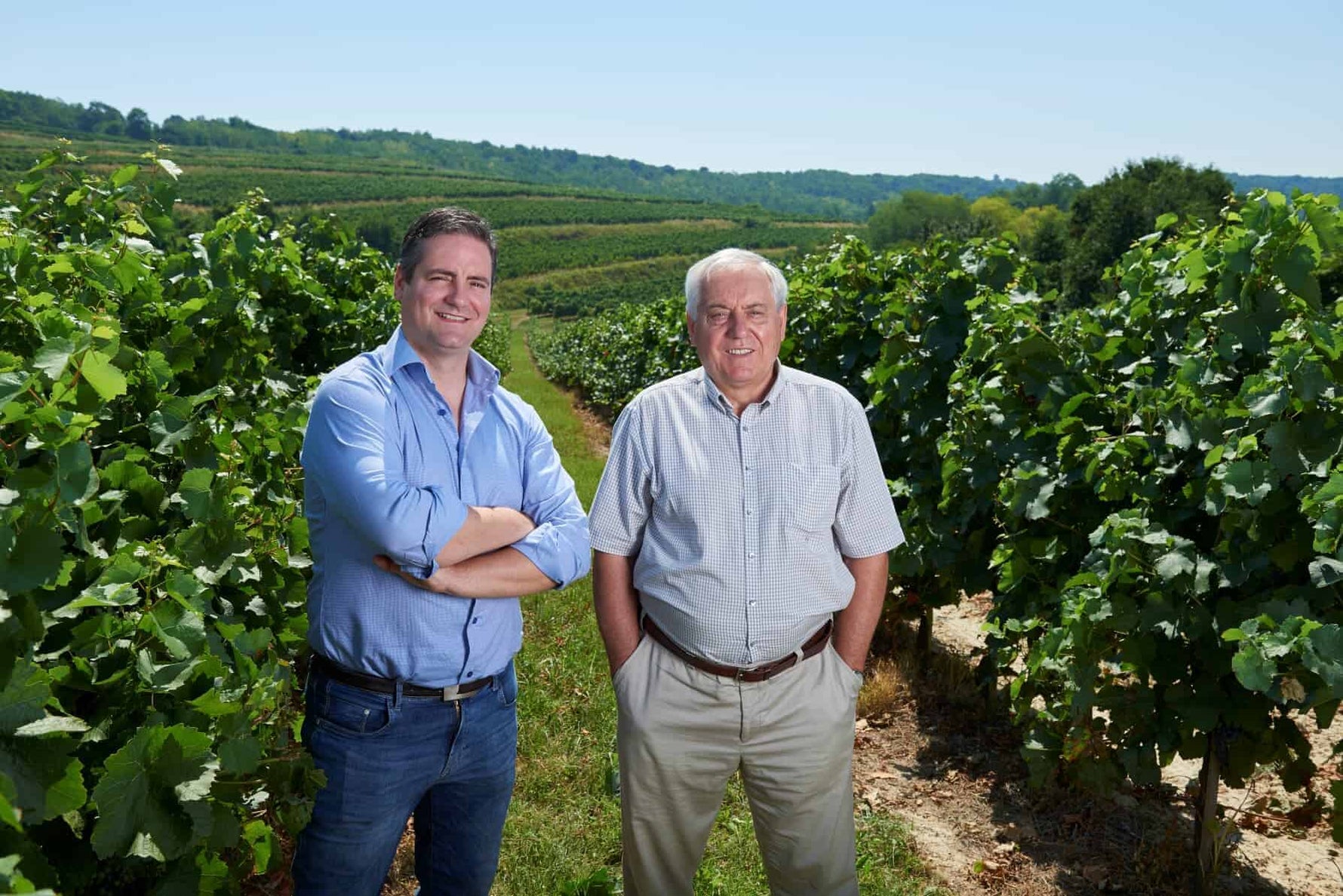 Zwei Männer stehen in einem Weinberg zwischen Reihen von Weinreben. Der Mann links trägt ein hellblaues Hemd und Jeans, der Mann rechts ein weißes Hemd und beige Hosen.
