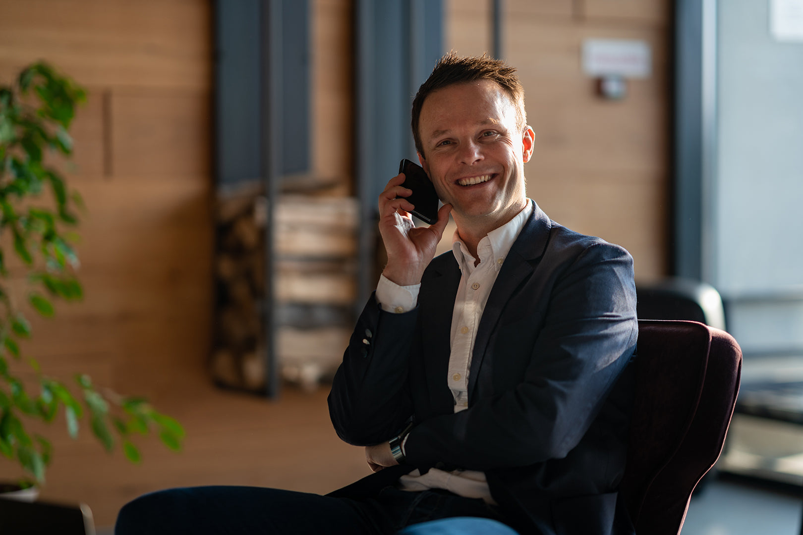 Mann in dunklem Anzug und weißem Hemd sitzt auf Stuhl und hält ein Smartphone ans Ohr, Hintergrund mit Holzpaneelen und Pflanze.