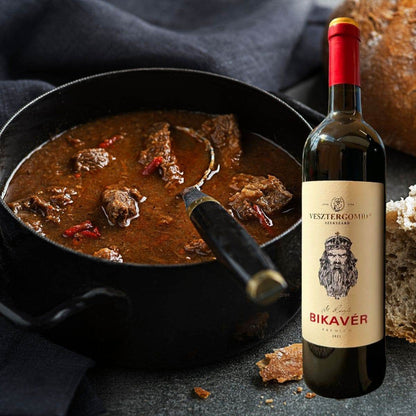 Schwarzer Topf mit ungarischem Gulasch und einem Löffel, daneben eine Flasche Vesztergombi St. László Bikavér Rotwein und Brot.