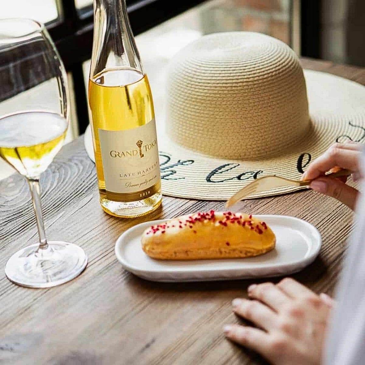 Flasche und Glas Grand Tokaji Late Harvest Weißwein, Eclair mit weißer Glasur und roten Streuseln auf weißem Teller, Strohhut