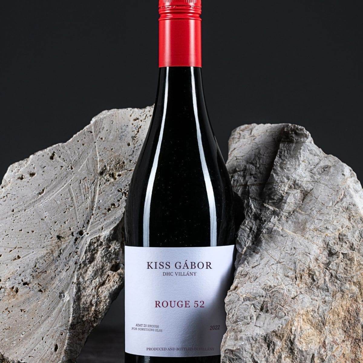 Flasche Kiss Gábor Rouge 52 Rotwein mit rotem Verschluss zwischen zwei grauen Steinen vor schwarzem Hintergrund