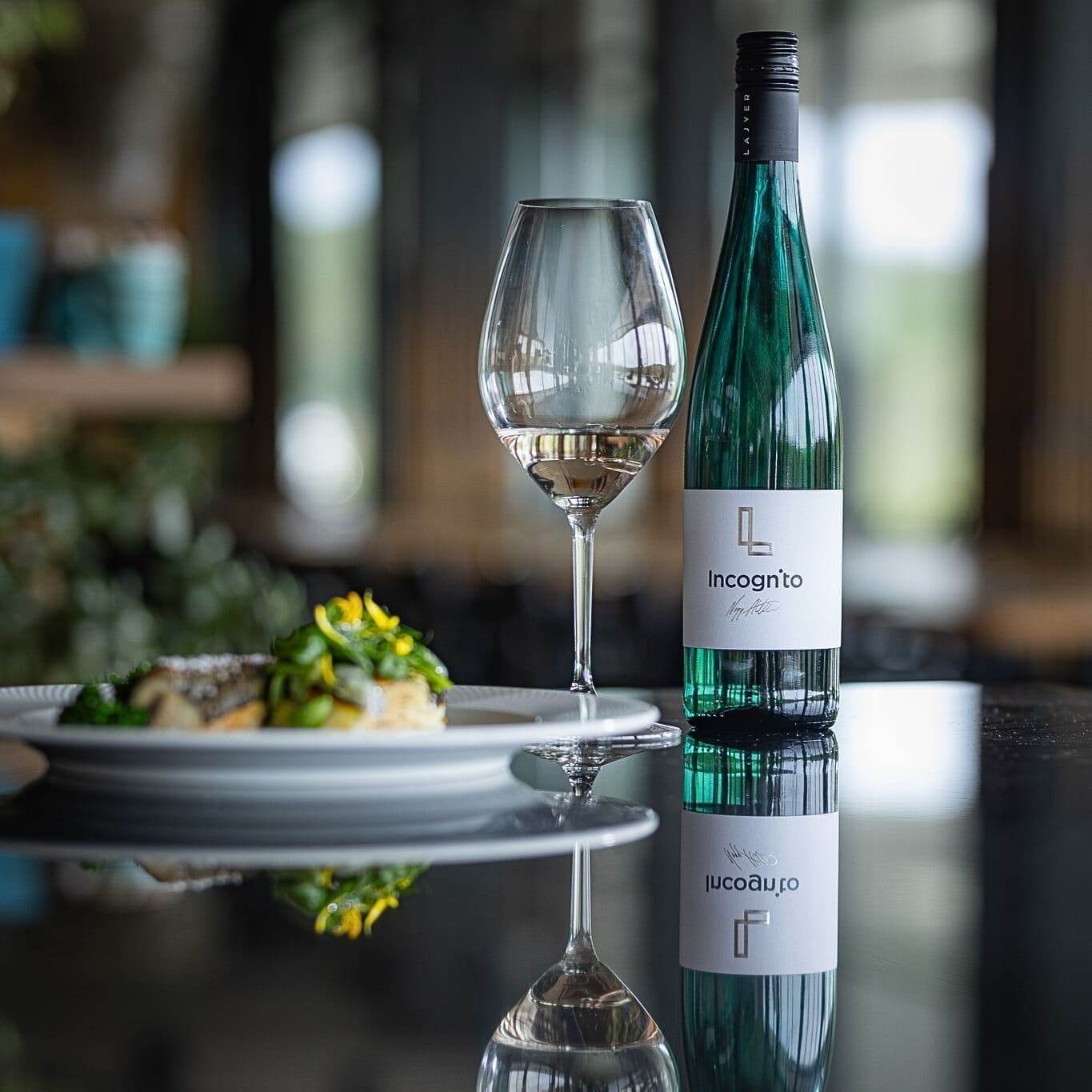 Grüner Weinflasche mit Etikett 'Incognito' neben einem Weinglas mit Weißwein und einem Teller mit dekoriertem Essen auf schwarzem Spiegeltisch.
