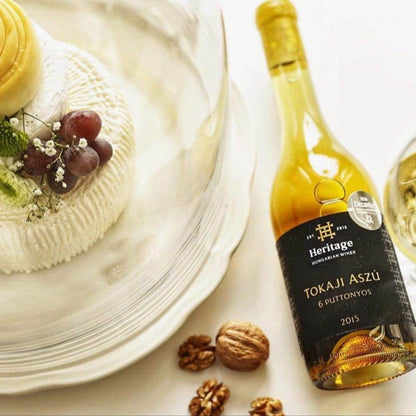 Flasche Heritage Tokaji Aszú 2015 Weißwein neben Glas mit Wein, Käse unter Glasglocke mit Trauben und Walnüssen