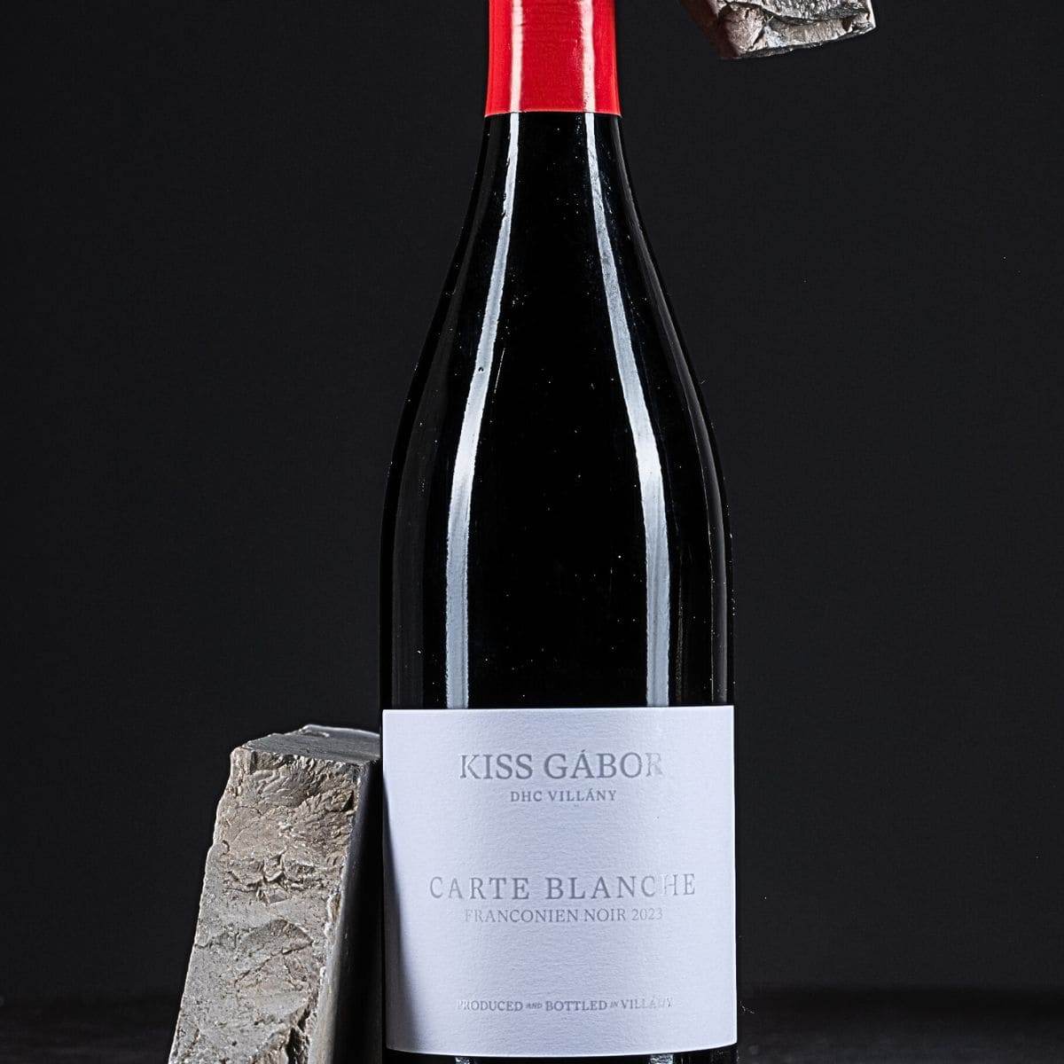 Flasche Kiss Gábor Carte Blanche Franconien Noir 2023 mit rotem Verschluss, umgeben von zwei grauen Steinen