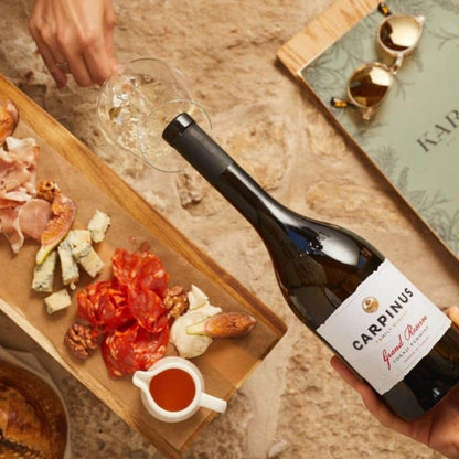 Hand hält eine Flasche Carpinus Grand Reserve Wein neben einer Holzplatte mit verschiedenen Käsesorten, Wurst und Walnüssen