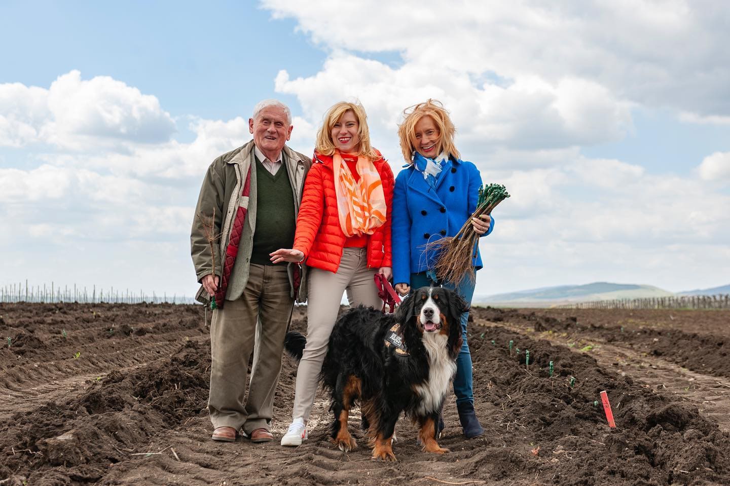 Drei Personen stehen auf einem Feld mit brauner Erde, umgeben von Pflanzenreihen. Ein schwarzer Hund sitzt vor ihnen. Die Person links trägt einen grauen Anzug, die mittlere Person eine orange Jacke und die rechte Person einen blauen Mantel.