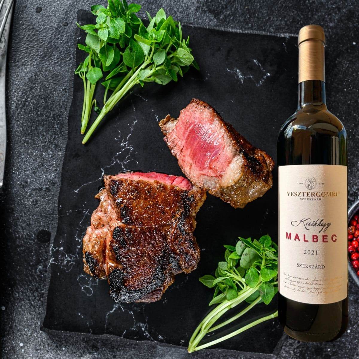 Gegrilltes, medium-rare Steak auf schwarzem Stein mit frischen grünen Kräutern neben einer Flasche Vesztergombi Kerékhegyi Malbec.