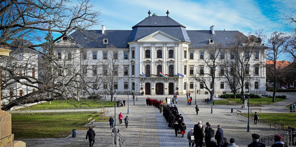 Das Gebäude der Eszterházy Károly Universität in Eger, mit einer symmetrischen Fassade und einem großen zentralen Eingang. Eine Gruppe von Personen in schwarzer Kleidung steht in einer Schlange auf der Treppe.