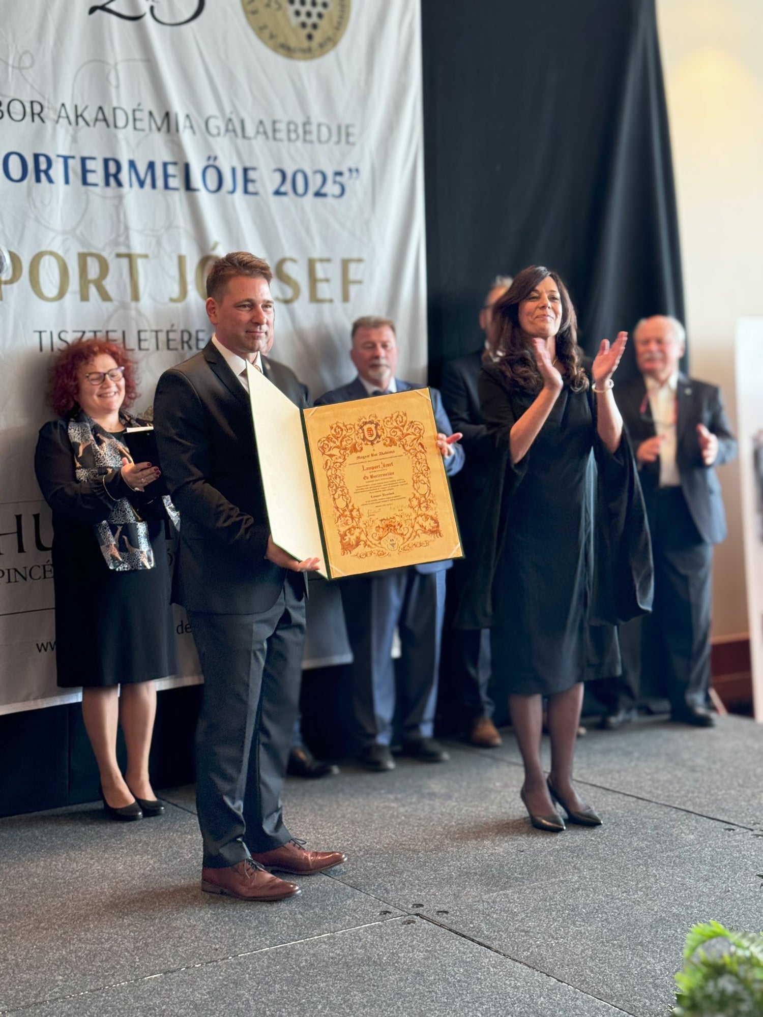 Mann in Anzug hält gerahmte Urkunde, umgeben von applaudierenden Personen auf Bühne vor Banner mit Text