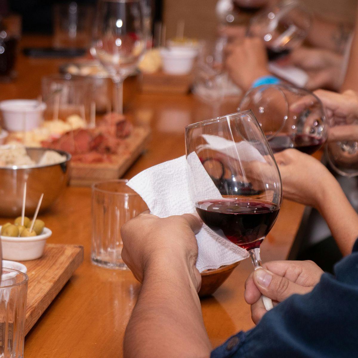 Eine Hand hält ein Weinglas mit rotem Wein, während eine andere Hand eine Serviette hält. Im Hintergrund sind mehrere Gläser und eine Platte mit Speisen zu sehen.