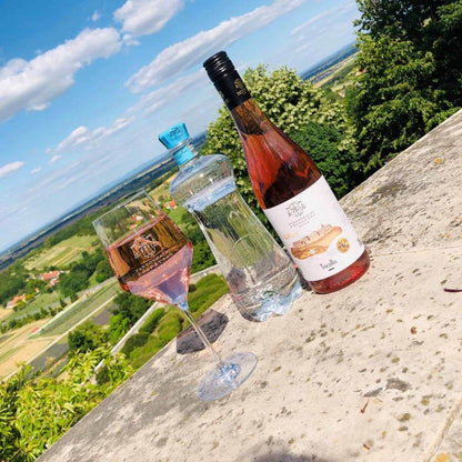 Flasche Roséwein, Flasche Mineralwasser und gefülltes Weinglas auf einer Steinmauer mit Blick auf eine grüne Landschaft und blauen Himmel