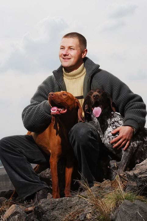 Mann sitzt auf Felsen mit zwei Hunden. Ein Hund ist rotbraun, der andere ist schwarz-weiß gefleckt. Beide Hunde haben ihre Zungen herausgestreckt.