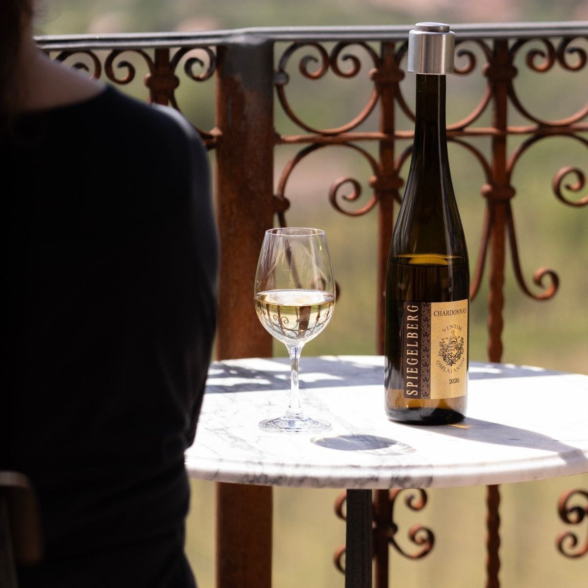 Flasche Spiegelberg Chardonnay 2020 und gefülltes Weinglas auf rundem Marmortisch, Person im schwarzen Oberteil sitzt daneben