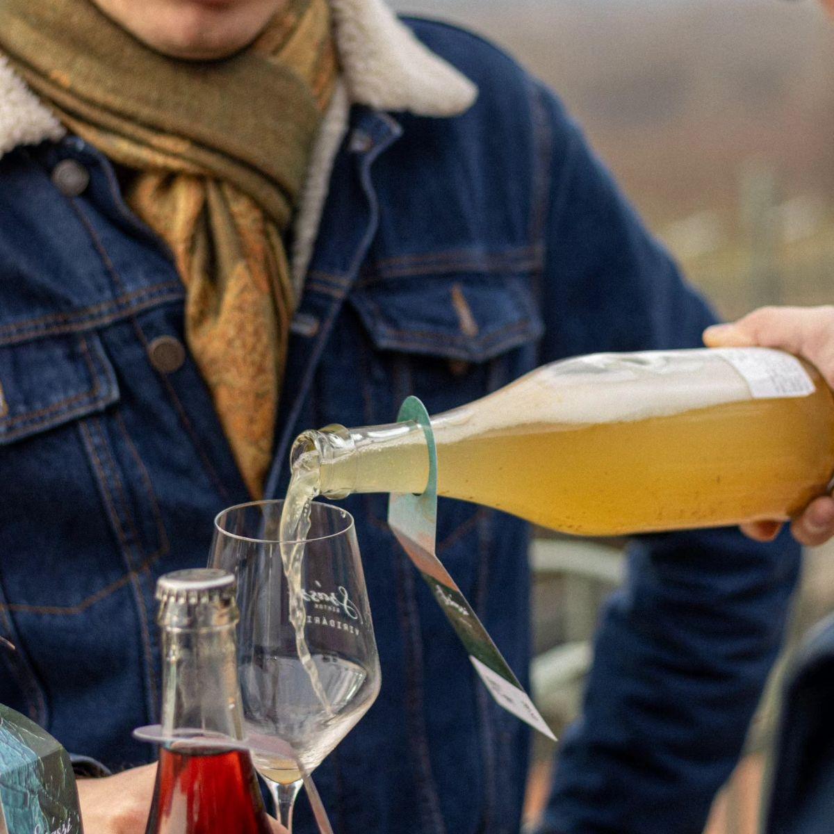 Person in Jeansjacke und Schal gießt ein helles Getränk aus einer Flasche in ein Weinglas