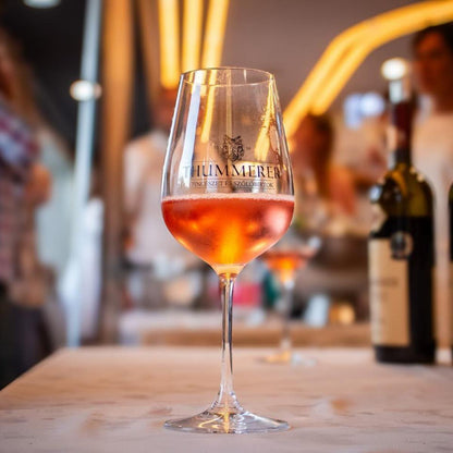 Ein Weinglas mit roséfarbenem Getränk beschriftet mit 'THUMMERER, steht auf einem Tisch. Im Hintergrund verschwommene Flasche