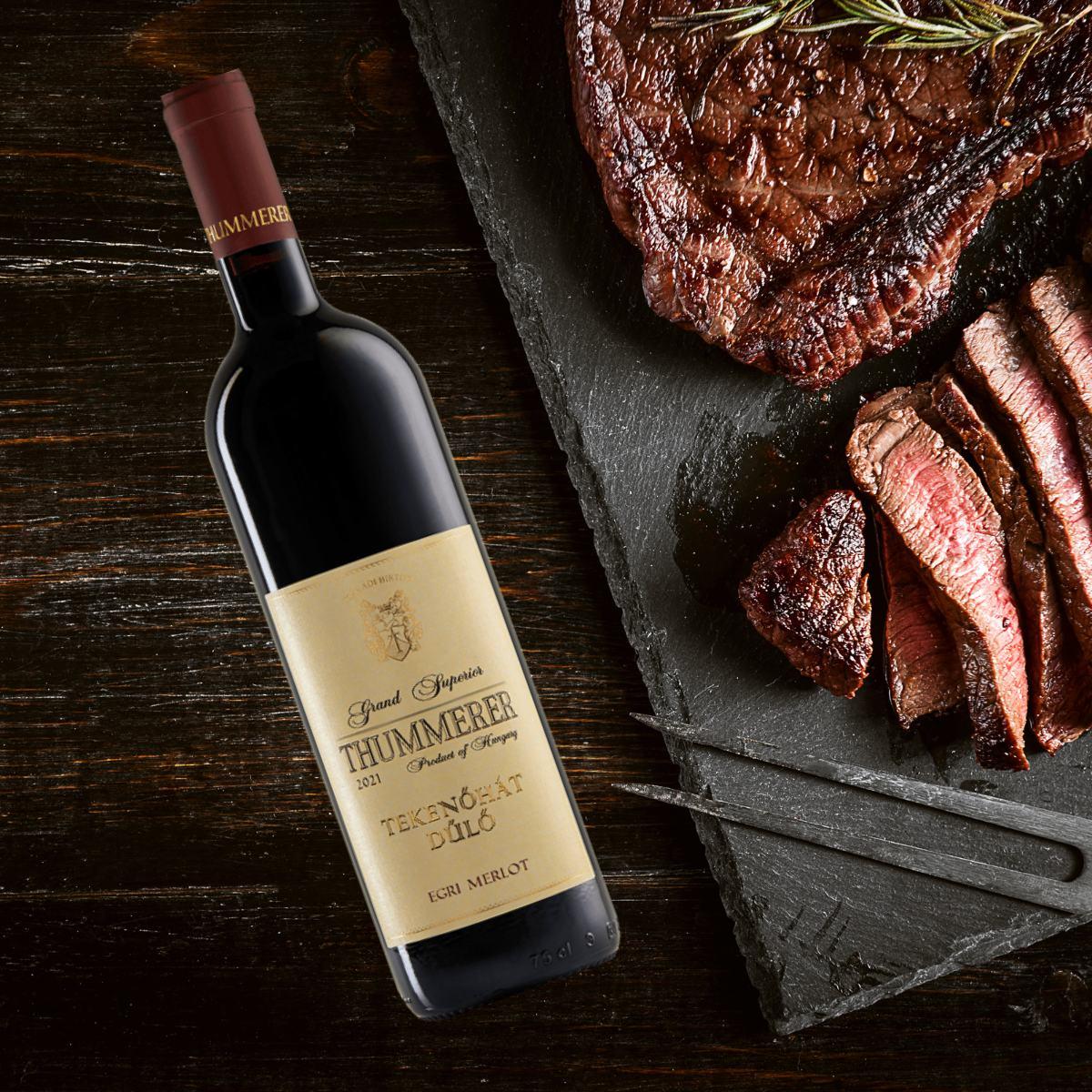 Eine Flasche Thummerer Tekenoháti Merlot 2021 steht neben einem geschnittenen Stück Rindersteak auf einem schwarzen Brett.