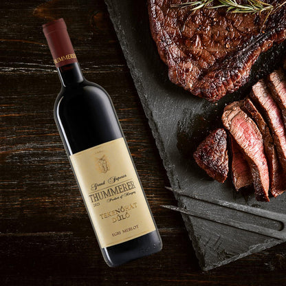 Eine Flasche Thummerer Tekenoháti Merlot 2021 steht neben einem geschnittenen Stück Rindersteak auf einem schwarzen Brett.